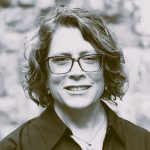 smiling woman with wavy, chin length hair wearing eyeglasses, a necklace and a dark, collared blouse.