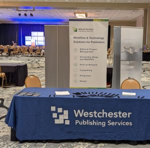 conference table with blue tablecloth on top of it, and pens and notebooks. Behind the table are two chairs and a rollup banner featuring services.