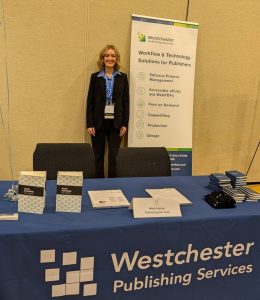 young woman with short blonde hair standing behind a conference table and next to a rollup banner.